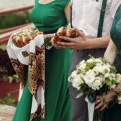 Wunderschöne Brautjungfern in stilvollen grünen Kleidern mit weißen Blumensträußen und traditionellen Broten in den Händen neben einem gutaussehenden Trauzeugen bei einer Hochzeitszeremonie im Freien, Nahaufnahme der Hände